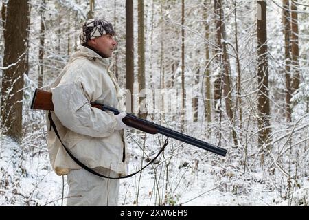 un chasseur en camouflage blanc avec un fusil dans les mains se tient dans une forêt hivernale enneigée pendant une chasse Banque D'Images