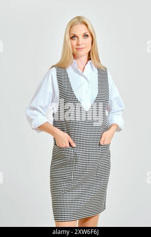 Femme mignonne en chemise blanche et robe de genou sans manches noir et blanc à carreaux posant sur fond gris clair Banque D'Images