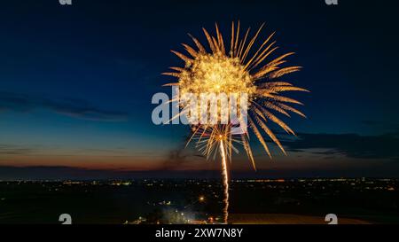 Des feux d'artifice dorés explosent dans le ciel nocturne, formant Un grand éclat radieux au-dessus des lumières lointaines de la ville et Une foule rassemblée, avec l'horizon crépusculaire rehaussant l'atmosphère festive. Banque D'Images