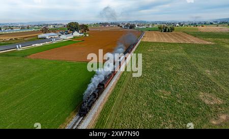 Un train à vapeur se déplace le long des voies entourées de champs verdoyants et de terres agricoles sous un ciel nuageux. Banque D'Images