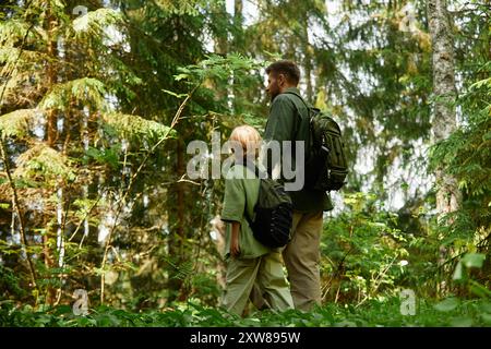 Deux individus avec des sacs à dos naviguant à travers la forêt verdoyante au milieu de la lumière du soleil filtrant à travers les arbres. Homme de grande taille et personnage aux cheveux blonds plus courts exprimant le sens de l'exploration Banque D'Images