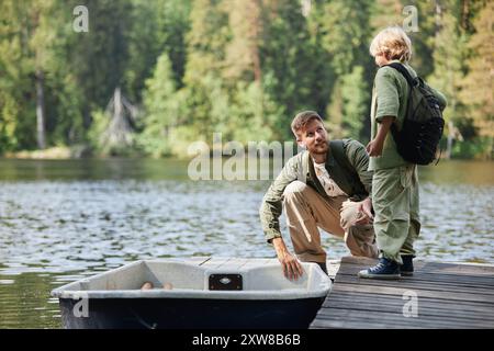 Père agenouillé sur le quai tout en parlant à son fils au bord du lac entouré d'arbres, tous deux vêtus de vêtements de plein air décontractés portant des engins de pêche Banque D'Images