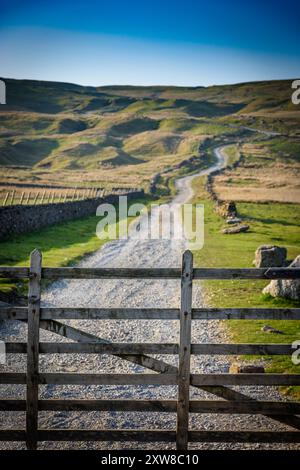 Une porte en bois est ouverte, révélant une piste de gravier qui monte à travers des collines vertes et brunes sous un ciel bleu clair. L'herbe luxuriante borde le p Banque D'Images