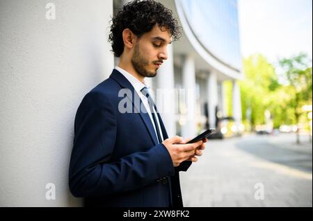 Jeune homme professionnel en costume engagé avec son téléphone à côté d'un immeuble de bureaux Banque D'Images