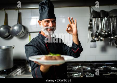 Le chef souriant tient une assiette de nourriture et fait le signe OK avec sa main. Il porte un uniforme de chef noir et un chapeau Banque D'Images