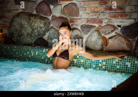 Femme se détend dans un bain à remous spa, souriant paisiblement. Les murs de pierre créent une ambiance tranquille. Cette image capture le bien-être et la beauté dans un cadre serein Banque D'Images