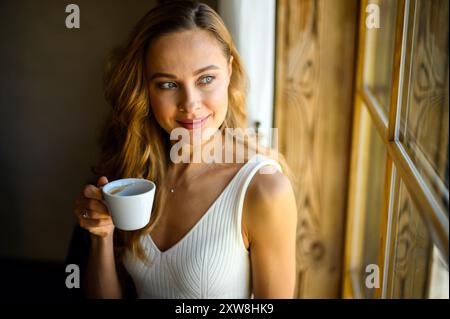 Jeune femme dans une robe blanche profite d'un moment paisible dans un café confortable, regardant par une fenêtre avec une tasse de café, souriant soigneusement Banque D'Images