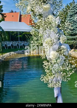 Arche festive décorée de fleurs blanches, construite à côté d'un petit lac de jardin. Herbe vert vif, plantes et arbres. Préparé pour un mariage et un jardin Banque D'Images