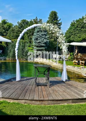 Arche festive décorée de fleurs blanches, construite à côté d'un petit lac de jardin. Herbe vert vif, plantes et arbres. Préparé pour un mariage et un jardin Banque D'Images