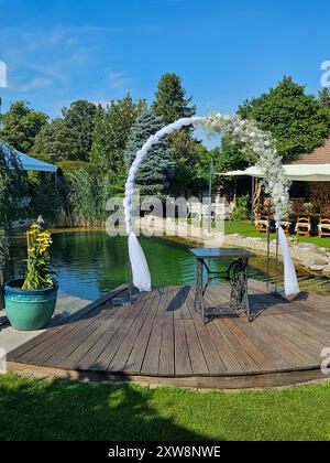 Arche festive décorée de fleurs blanches, construite à côté d'un petit lac de jardin. Herbe vert vif, plantes et arbres. Préparé pour un mariage et un jardin Banque D'Images