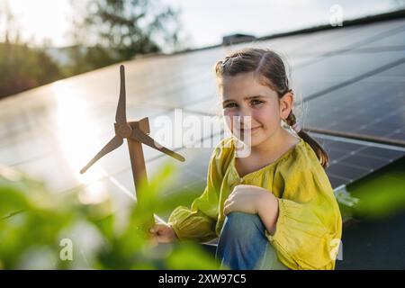 Fille mignonne sur le toit avec des panneaux solaires, tenant le modèle de l'éolienne. Installation solaire ou photovoltaïque sur le toit. Un avenir durable pour la prochaine génération. Banque D'Images