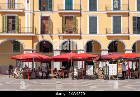 Bâtiment coloré à Palma avec des volets en bois fermés contrastés, donnant sur une place remplie de restaurants en plein air et de parasols rouges vibrants. Banque D'Images