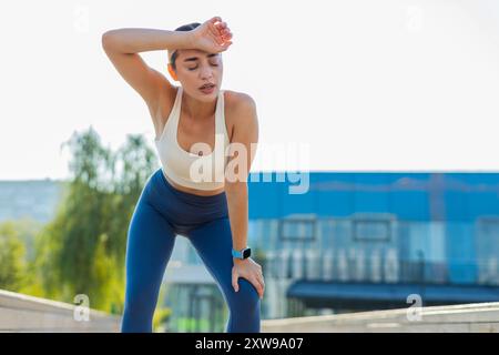 La femme de coureur athlétique épuisée regarde le bracelet de remise en forme de l'appareil de montre intelligente vérifiant le temps fréquence cardiaque, pouls, SpO2 après le jogging. Jeune femme fatiguée travaillant cardio dehors dans le parc urbain ensoleillé de la ville Banque D'Images