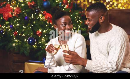 Couple afro romantique buvant du champagne près de l'arbre de Noël, célébrant les vacances Banque D'Images