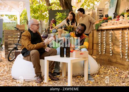 Vendeur de personnes âgées caucasiennes et homme noir clinquant des verres de vin biologique au festival local de la foire des récoltes. Personnes multiraciales appréciant au marché des agriculteurs, deux hommes buvant naturellement fait bio produit. Banque D'Images