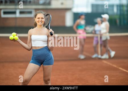 Jeune fille joyeuse avec raquette et balles de tennis debout sur un court extérieur Banque D'Images