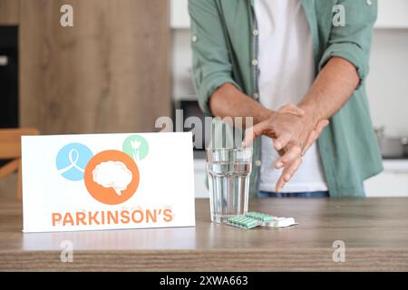 Papier avec le mot PARKINSON'S, pilules et homme senior prenant un verre D'eau de la table dans la cuisine, gros plan Banque D'Images