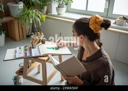 Artiste de jeune fille créative focalisée redessinant avec des esquisses d'aquarelles du bloc-notes dans le studio d'atelier d'art Banque D'Images