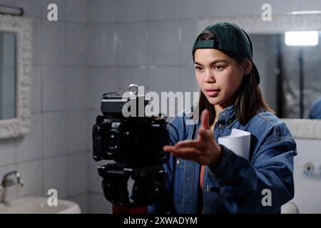 Femme ajustant l'appareil photo avec une lampe de poche dans la salle de bain carrelée tout en tenant des papiers, apparaissant concentré sur la configuration précise, portant une veste en Jean et une casquette arrière Banque D'Images