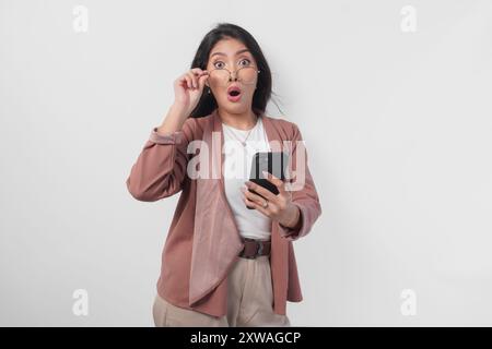 Femme d'affaires asiatique surprise dans des lunettes regardant des nouvelles choquantes sur son téléphone, isolée par fond blanc. Banque D'Images