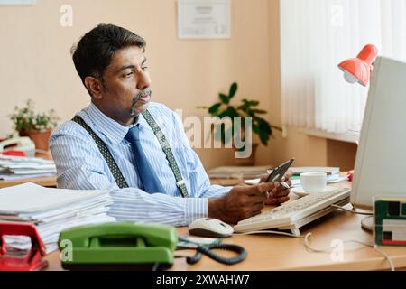 Homme d'affaires assis au bureau tenant smartphone dans une main tout en regardant soigneusement l'écran d'ordinateur avec des plantes et des documents en arrière-plan Banque D'Images