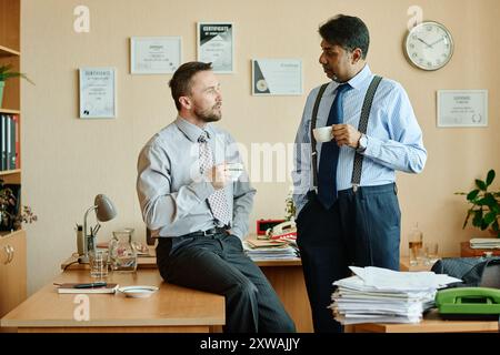 Deux hommes d'affaires dans le cadre de bureau tenant des tasses et ayant la conversation sur les stratégies d'affaires tout en se tenant debout et assis près des bureaux couverts de documents et de fournitures de bureau Banque D'Images