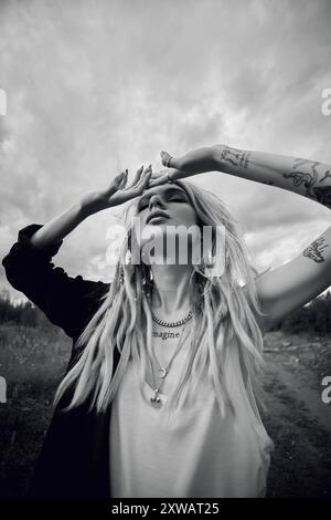 Une femme avec de longs dreadlocks blonds et plusieurs colliers se tient dans un champ, regardant le ciel nuageux. Ses mains sont levées, protégeant ses yeux fr Banque D'Images