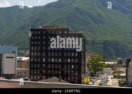 Bolzano, Italie - 8 juin 2023 : un immeuble de bureaux moderne Enzian bureau avec une façade en verre se dresse sur un fond de montagnes verdoyantes et verdoyantes. Voiture Banque D'Images