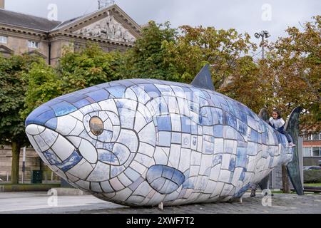 Belfast, Royaume-Uni - 17 août 2024 : enfant jouant sur la queue d'une grande sculpture de poisson bleu et blanc le long des rives de la rivière Lagan dans le centre-ville. Banque D'Images