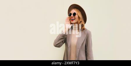 Portrait d'un beau modèle jeune femme souriant portant un chapeau rond brun et un manteau sur fond beige Banque D'Images