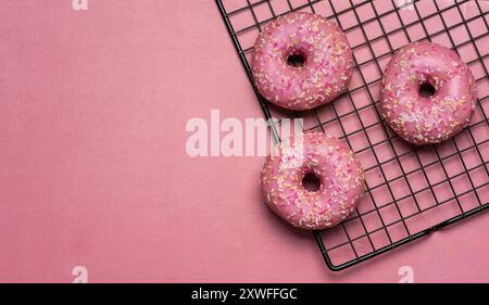 Trois beignets roses givrés avec des saupoudres multicolores reposent partiellement sur une grille de refroidissement, placée sur un fond rose, dégageant un délicieux et invitant Banque D'Images