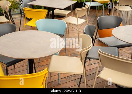 Une variété de chaises vides colorées et de tables rondes disposées sur un patio extérieur en bois. Le coin salon moderne est prêt pour les visiteurs, parfait pour CAF Banque D'Images