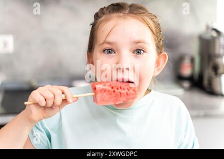 Une petite fille mange une pastèque sur un bâton sur le fond de la cuisine, elle se tient debout en regardant le cadre habillé d'un T-shirt turquoise. Élevé Banque D'Images