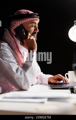 Concentré sur la conversation téléphonique est un homme d'affaires déterminé portant des vêtements islamiques et vérifiant l'ordinateur portable. Vue latérale d'un musulman en train de faire plusieurs tâches, à l'aide de son ordinateur personnel et de son appareil mobile. Banque D'Images