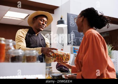 Une femme afro-américaine discute des achats respectueux de l'environnement avec le commerçant à la caisse. Une cliente donnant le pot de pâtes au vendeur masculin à la caisse. Banque D'Images