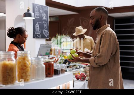 Vendeur féminin afro-américain aidant les clients sur le marché moderne. Produits frais, biologiques et écologiques dans des pots en verre, avec emballage en papier. Mise au point sélective sur le comptoir de caisse. Banque D'Images