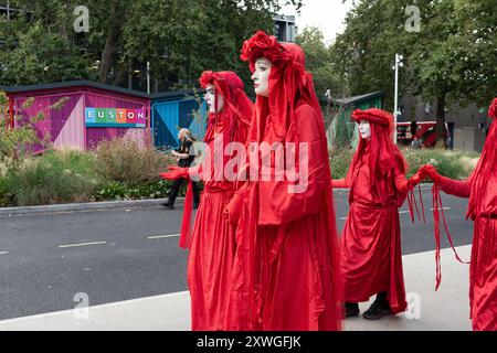 Londres, Royaume-Uni. 19 août 2024. La troupe mime de l'activiste Red Rebel d'extinction Rebellion traverse les jardins d'Euston Square, en face de la gare d'Euston, pour protester silencieusement contre les impacts environnementaux du projet ferroviaire HS2, y compris la perte d'arbres et d'espaces verts. La zone, maintenant une station de taxis, était autrefois un parc public et a été occupée par des manifestants dans des camps d'arbres et des tunnels en 2021 dans le but de la sauver. Le projet, dont l'avenir est incertain, a dépassé largement le budget et a provoqué des bouleversements massifs dans la région. Crédit : Ron Fassbender/Alamy Live News Banque D'Images