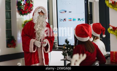 Superviseur habillé en Père Noël pendant la saison de Noël tenant une réunion d'équipe, montrant les données de l'entreprise sur l'écran numérique. Chef d'équipe dans le lieu de travail décoré de Noël discutant avec les employés Banque D'Images