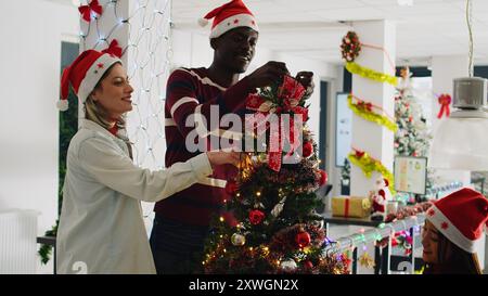 Les collègues qui décorent le sapin de Noël avec des boules et des guirlandes au travail à Noël ornent le lieu de travail. Les membres du personnel s'amusent ensemble dans un bureau décoré festif pendant la saison des vacances d'hiver Banque D'Images