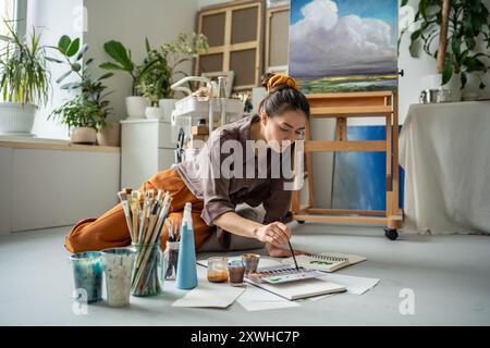 Inspirée jeune fille créative illustrateur peinture avec des aquarelles dans le carnet de croquis sur le sol dans l'atelier d'art. Banque D'Images