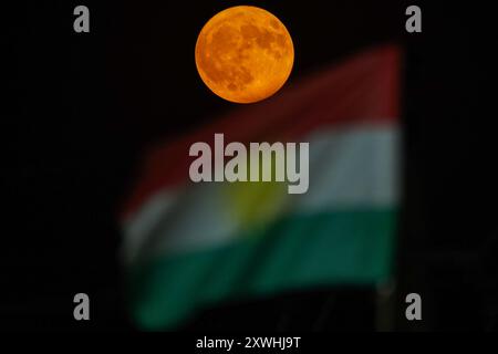 Duhok, Irak. 19 août 2024. La super lune se lève derrière le drapeau de la région du Kurdistan dans le ciel de la ville de Duhok, au nord de l'Irak. Crédit : SOPA images Limited/Alamy Live News Banque D'Images