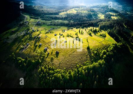 Vue aérienne de la vallée ensoleillée, parsemée de petites maisons et de routes sinueuses, entourée de forêts denses. La lumière dorée projette de longues ombres. Un paysage dynamique s'étend, se fondant dans des collines boisées. Banque D'Images