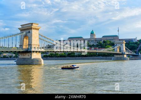 Budapest, Hongrie - 7 juillet 2024 : Pont à chaînes enjambant le Danube avec le château de Buda en arrière-plan, avec un petit bateau naviguant sous un p Banque D'Images