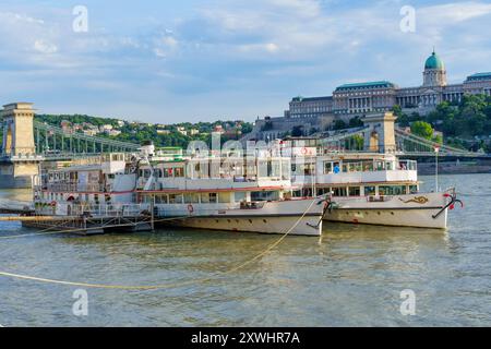 Budapest, Hongrie - 7 juillet 2024 : bateaux de tourisme amarrés sur le Danube avec le pont des chaînes et le château de Buda visibles en arrière-plan. Banque D'Images