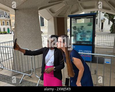 Londres, Royaume-Uni. 14 août 2024. La boîte de sentinelle de la police avec l'illustration de Banksy représentant des piranhas, qui était à l'origine sur Ludgate Hill, a maintenant été déplacée à Guildhall par les fonctionnaires de la City of London. Deux femmes prenant des selfies sur leur téléphone portable. Banque D'Images