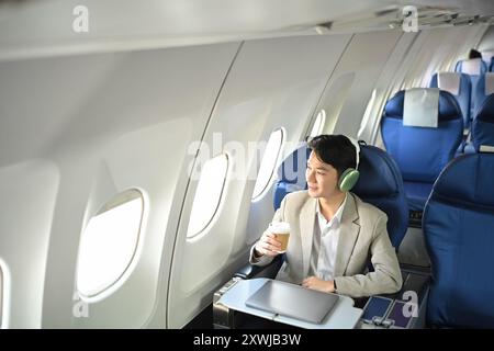 Agréable jeune homme d'affaires avec une tasse de café écoutant de la musique dans des écouteurs assis dans l'avion pendant un vol Banque D'Images