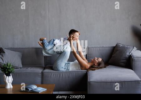 la jeune mère tient son petit fils dans ses bras et le soulève à la maison, le garçon de 2 ans s'amuse et joue avec son parent sur le canapé, la femme est rai Banque D'Images