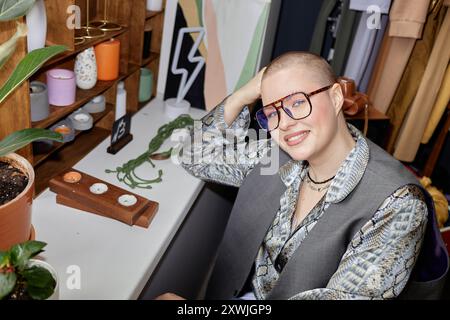 Portrait tourné avec flash de l'appareil photo de la jeune femme à la mode habillée dans le style vintage posant à côté de la vitrine avec des bougies et souriant tout en regardant la caméra à la boutique de seconde main, espace de copie Banque D'Images