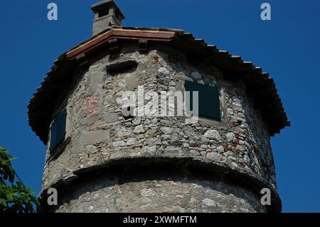 Une cheminée traditionnelle à persiennes s’élève au-dessus du toit en tuiles de terre cuite de cette tour circulaire en pierre aux volets en bois vert à Cassone di Malcesine, un village sur la rive est du lac de Garde en Vénétie, en Italie. La tour est connue sous le nom de la 'Toresela'. Banque D'Images