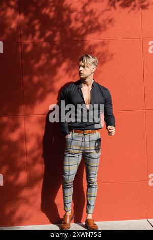 Un bel homme blond dans une chemise noire et un pantalon à carreaux se penche contre un mur rouge Banque D'Images